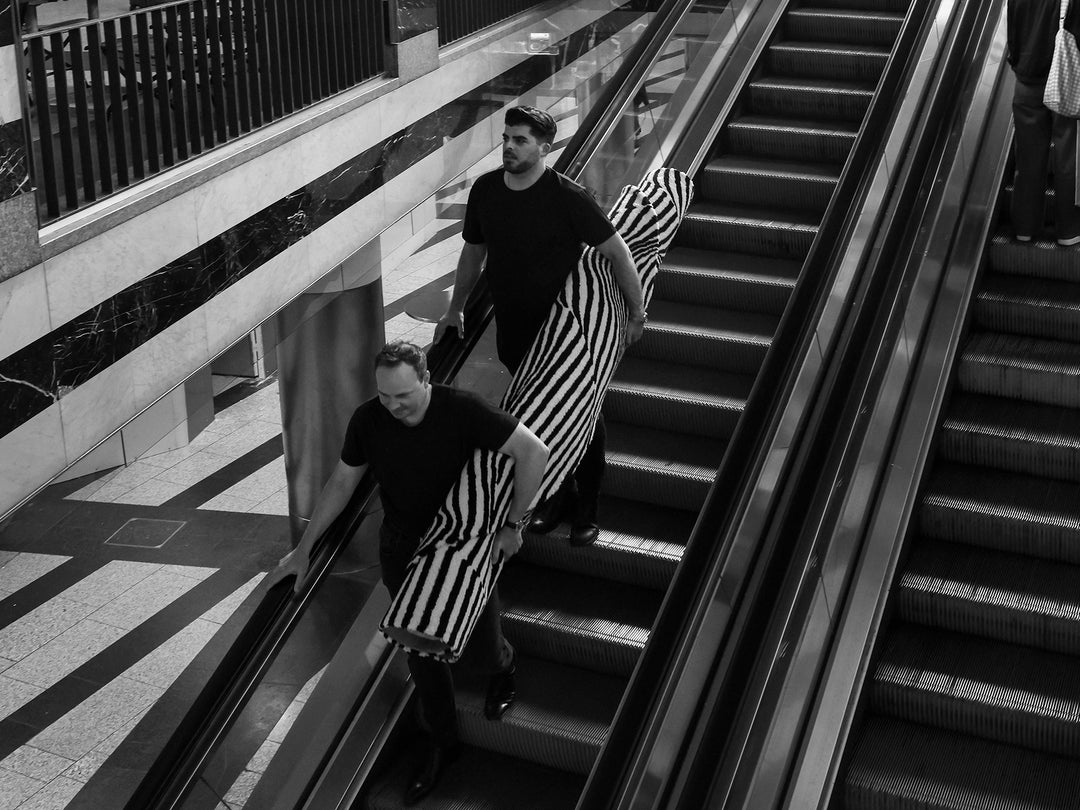 Schwarzweissaufnahme mit Personen und Teppichrollen auf einer Rolltreppe